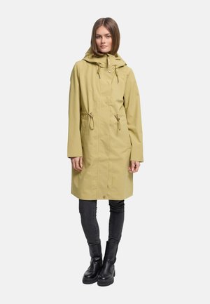 Žena oblečená v svetloolivovom pršiplášti s kapucňou po kolená so sťahovacími šnúrkami, čiernych úzkych džínsoch a čiernych členkových topánkach na jednoduchom pozadí.