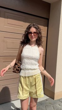 Halter-top i vitt med texturerade volanger, kombinerad med gula träningsshorts med vita sidostråkar och logotyp. Brun axelväska.