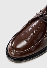 Brun laklædersko med rundet tå, glat overflade og kontrasterende sort sål. Har snørebånd og syningsdetaljer.