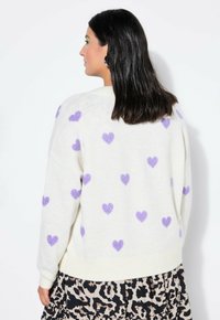 Kremowy sweter z rozrzuconymi fioletowymi wzorami serc, o luźnym kroju, z ściągaczem na dole i długimi rękawami, noszony na wzorzystej spódnicy.