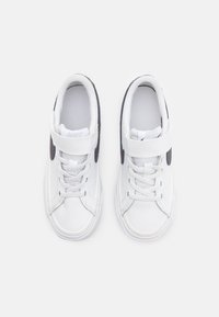 Ténis brancos com parte superior de couro liso, bico redondo e detalhes pretos nas laterais. Inclui atacadores e uma fita aderente para fecho.