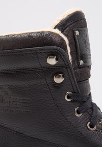 Bota de cuero negra con un forro de felpa beige, acentos metálicos plateados, ojales reforzados y detalle de logo embossado en el lateral.