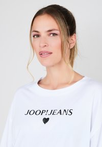 Weißes, übergroßes T-Shirt mit rundem Ausschnitt, das mit schwarzem "JOOP!JEANS"-Text und Herzgrafik gestaltet ist. Weicher Stoff mit glatter Textur.