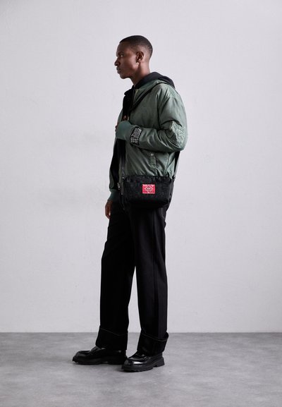Grüne Bomberjacke mit strukturierten Ärmeln, schwarze Umhängetasche mit rotem Akzent, weit geschnittene schwarze Hose und schwarze klobige Schuhe.