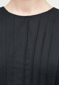 Κοντινή λήψη μαύρου πλισέ υφάσματος που δείχνει το άνω μέρος του στήθους και τον λαιμό ενός ατόμου που φοράει ρούχο με στρογγυλή λαιμόκοψη.