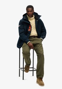 Giacca isolante blu navy da uomo con logo arancione, maglione lavorato a maglia beige, pantaloni cargo verde oliva e stivali color beige, seduto su uno sgabello contro uno sfondo bianco.