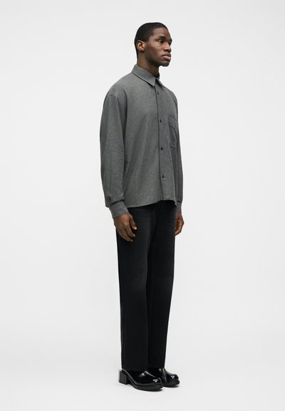 Άνδρας στέκεται γυρισμένος προς τα δεξιά, φοράει γκρι πουκάμισο με κουμπιά, μαύρο ίσιο παντελόνι και μαύρα παπούτσια με πάτο, μπροστά σε έναν απλό λευκό φόντο.