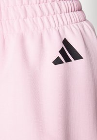 Gaiši rozā sporta bikses ar elastīgu jostu, kurās labajā pusē ir melns trīs svītru Adidas logotips. Gluda, mīksta tekstūra.