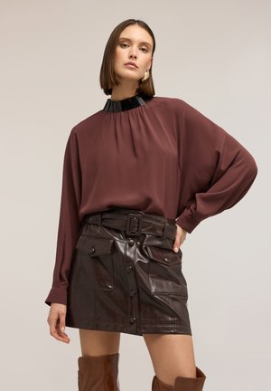 Kvinde med kort brunt hår iført en vinrød bluse med sort krave, sort lædernederdel med bælte og brune knæhøje støvler, står med hånden på hoften.