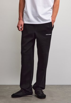 Man met zwarte gestructureerde broek met wit logo op de dij, wit overhemd en zwarte sneakers, staat op een grijze vloer.