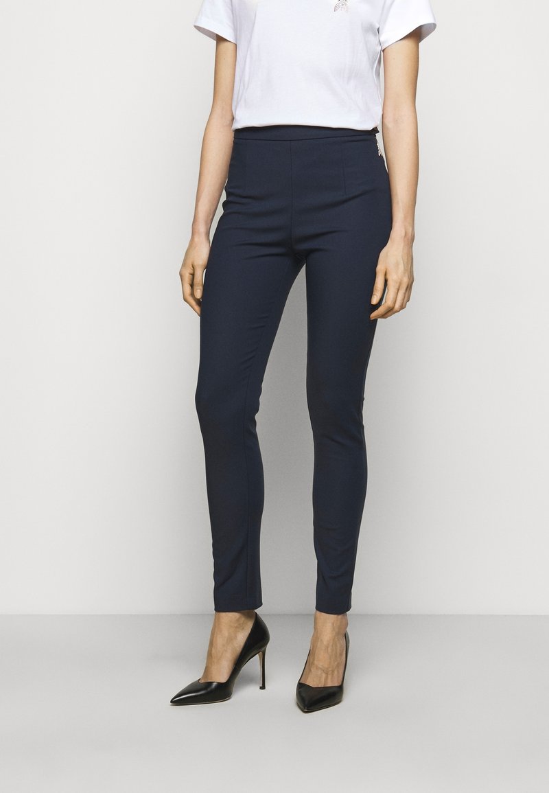 Marineblauwe, getailleerde broek met een gladde textuur, hoge taille en smalle pijpen, gecombineerd met een witte t-shirt met korte mouwen en zwarte hoge hakken.