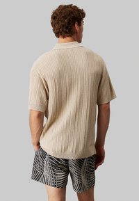Camisa polo de punto beige con mangas cortas, que presenta un tejido acanalado y un cuello. Combinada con pantalones cortos de patrón en blanco y negro.