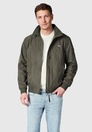 Uomo che indossa una giacca verde oliva con cerniera sopra un maglione bianco e jeans azzurri, in piedi con una mano in tasca davanti a uno sfondo semplice.