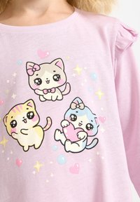 Light pink, short-sleeved shirt featuring cartoon kittens in various poses. The design includes pastel colors and hearts, with ruffled sleeve accents.