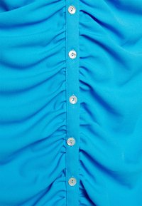 Peça de roupa abotoada em tecido canelado azul brilhante apresenta textura franzida na frente com botões brancos ao centro.