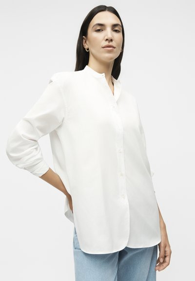 Γυναίκα με μακριά σκούρα μαλλιά φοράει άσπρο πουκάμισο με κουμπιά και ανοιχτό μπλε τζιν, ποζάροντας με το ένα χέρι στη μέση, μπροστά από απλό φόντο.