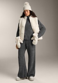 Sivý pletený relaxačný set s širokými nohavicami, doplnený krémovou fleecovou vestou a zodpovedajúcimi rukavicami. Celkový look dotvára svetlý pletený klobúk.