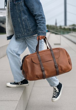 Person trägt Jeansjacke und Jeans, steigt auf eine Kante und trägt eine große braune Leder-Reisetasche mit dunklen Trägern.