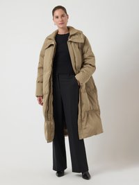 Lungo cappotto piumino beige con colletto alto e dettagli testurizzati, indossato sopra un maglione nero e pantaloni neri a vita alta.