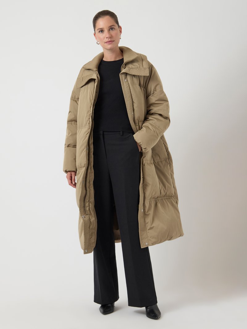Lungo cappotto piumino beige con colletto alto e dettagli testurizzati, indossato sopra un maglione nero e pantaloni neri a vita alta.