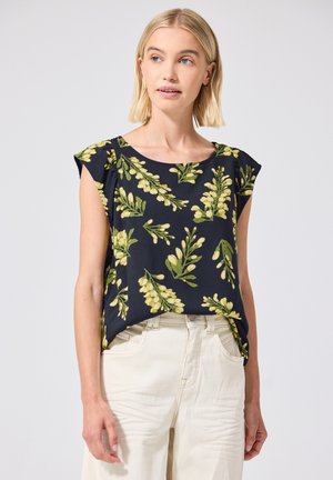 Kvinde iført en mørkeblå ærmeløs top med gult blomsterprint og høj taljede off-white bukser mod en ensfarvet baggrund.