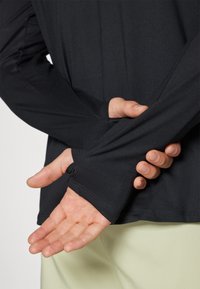 Schwarzes Langarmshirt mit glatter Textur, das Daumenlöcher an den Bündchen und ein tailliertes Design aufweist. Hellfarbige Hose ist darunter sichtbar.