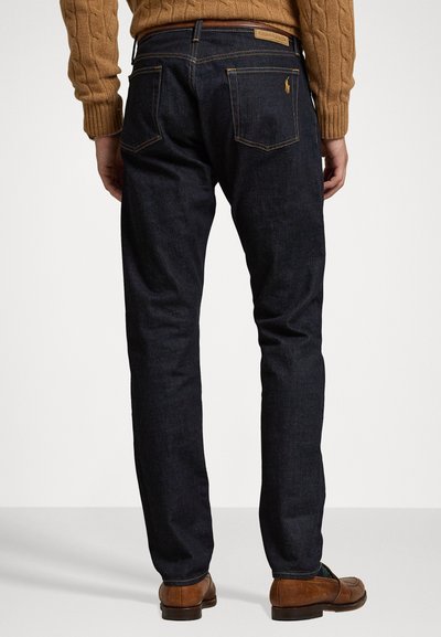 Mørkeblå denimbukser med rett passform, med kontrasterende gullsømmer og en liten logo på baklommen. Brune lærsko er synlige.