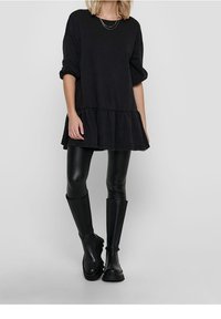 Kobieta w luźnej, czarnej sukience z długim rękawem i falbaniastym dołem, czarnych skórzanych legginsach i czarnych kozakach do kolan na jednolitym tle.
