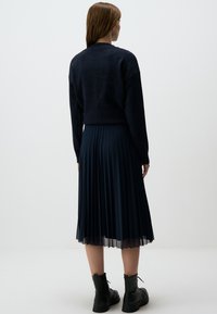 Gonna plissettata blu navy con dettagli trasparenti, abbinata a un maglione di lana scuro. Modella in piedi con stivali neri alla caviglia, vista dal retro.