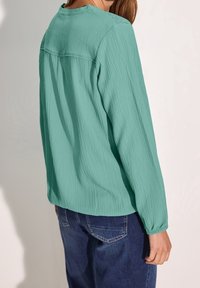 Blouse texturée bleu sarcelle clair, manches longues, col rond et poignets froncés, associée à un jean en denim bleu.