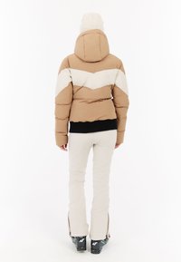 Pufová bunda v béžovej a bielej farbe s vzorom chevron, s kapucňou a čiernym elastickým lemom. Nosená s bielymi lyžiarskymi nohavicami a čižmami.