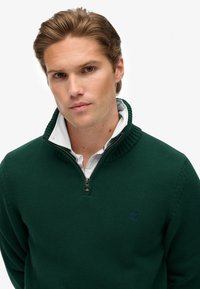 Suéter verde de punto con cuello alto, cierre de media cremallera y tejido texturizado. Lleva una camisa blanca debajo con cuello, que presenta un logo.