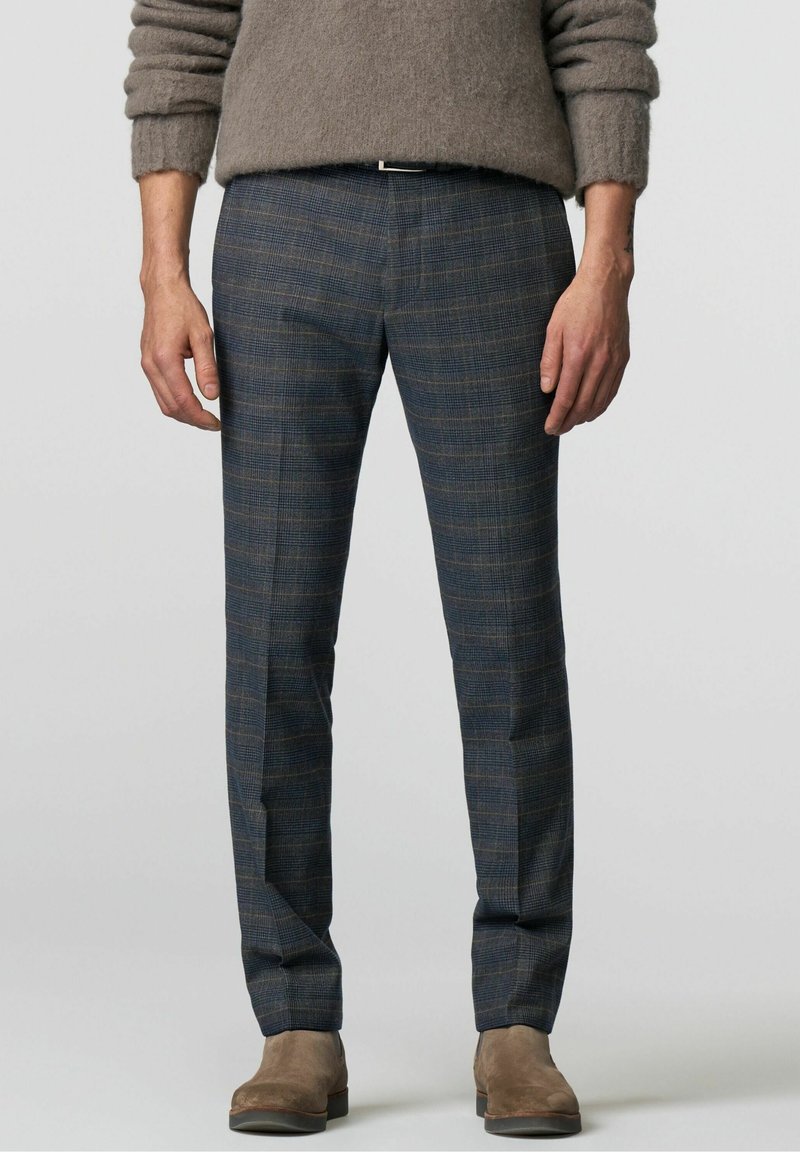 Homme portant un pantalon à carreaux gris slim, des bottes en daim beige et un pull duveteux taupe, debout les mains détendues le long du corps.