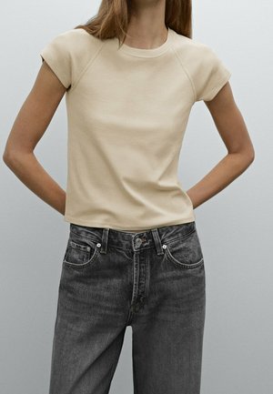 Personne mince portant un haut beige à manches courtes et un jean noir délavé taille haute, les mains reposant derrière le dos.