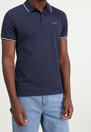 Mand iført navyblå polo med hvide kanter og lyseblå jeans mod en ensfarvet baggrund.
