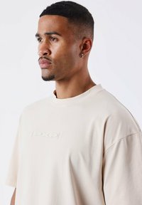 Camiseta de algodón beige de corte holgado con una textura suave, que presenta un logo texturizado "PXP" en el pecho y un cuello redondo.
