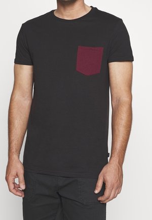 Mann trägt ein schwarzes Kurzarm-T-Shirt mit einer dunkelroten Brusttasche und schwarzen Hosen, steht vor einem schlichten Hintergrund.