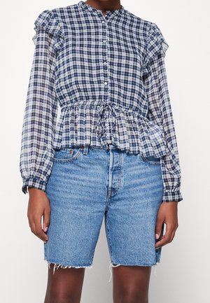Kvinde iført en langærmet blå og hvid ternet bluse bundet i taljen og blåt denimbukshorts i knælængde med frynset kant.