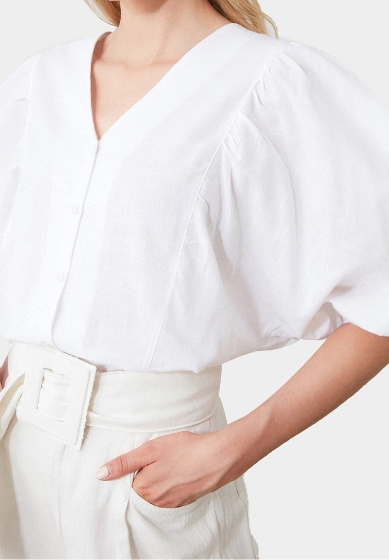 Blusa di lino bianca con maniche a sbuffo, scollatura a V profonda, bottoni frontali e tessuto testurizzato; abbinata a pantaloni crema a vita alta e cintura.