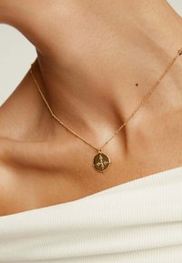 Collar de oro con un colgante ovalado que presenta un diseño geométrico, suspendido en una delicada cadena sobre una blusa blanca texturizada.