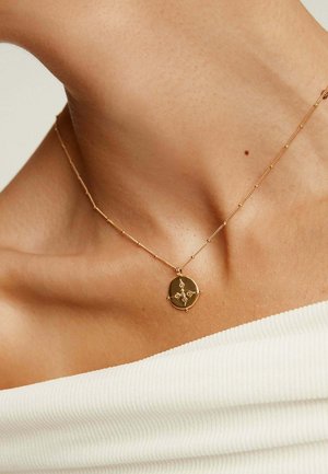 Collar de oro con un colgante ovalado que presenta un diseño geométrico, suspendido en una delicada cadena sobre una blusa blanca texturizada.