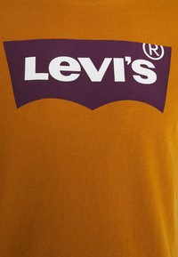 Pomarańczowa bawełniana koszulka z dużym, purpurowym logo Levi's nadrukowanym dużymi białymi literami wewnątrz zakrzywionego kształtu emblemu.
