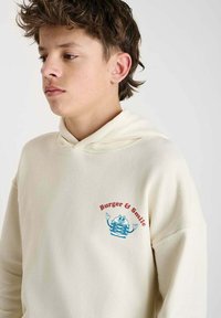 Crème-kleurige hoodie met een voorzak, voorzien van een blauw en rood graphic met de tekst "Burger & Smile" naast een hamburgerfiguur.