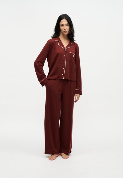 Femme debout pieds nus portant un pyjama bordeaux à manches longues avec passepoil blanc, une main dans la poche, sur un fond blanc uni.