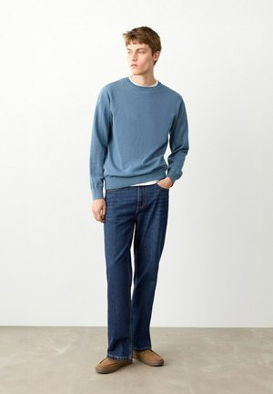 Giovane uomo in piedi, indossa un maglione azzurro chiaro, jeans blu scuro e scarpe marroni, guarda verso sinistra su uno sfondo bianco semplice.