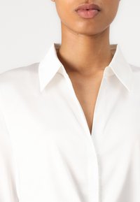 Camicia bianca con colletto classico, tessuto liscio e pattella con bottoni. Nessun motivo o decorazione visibile.