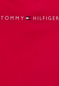 Rdeča tekstilna tkanina z gladko teksturo, ki vsebuje kontrastni beli in črni logotip pas z napisom "TOMMY HILFIGER" na vrhu.
