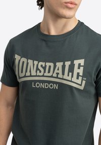 Tamsiai žalia medvilninė marškinė su dideliu smėlio spalvos "LONSDALE" logotipu ir žodžiu "LONDON" žemiau, su klasikiniu apvaliu kaklu.