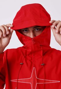 Persoon in een rode hoodie met een wit sterrenontwerp, die de capuchon aanpast en gedeeltelijk hun gezicht bedekt.
