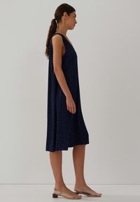 Frau im Profil, die ein ärmelloses, knielanges Kleid in marineblau mit kleinem Punkte-Muster und beigen, flachen Sandalen trägt, vor einem einfarbigen Hintergrund.